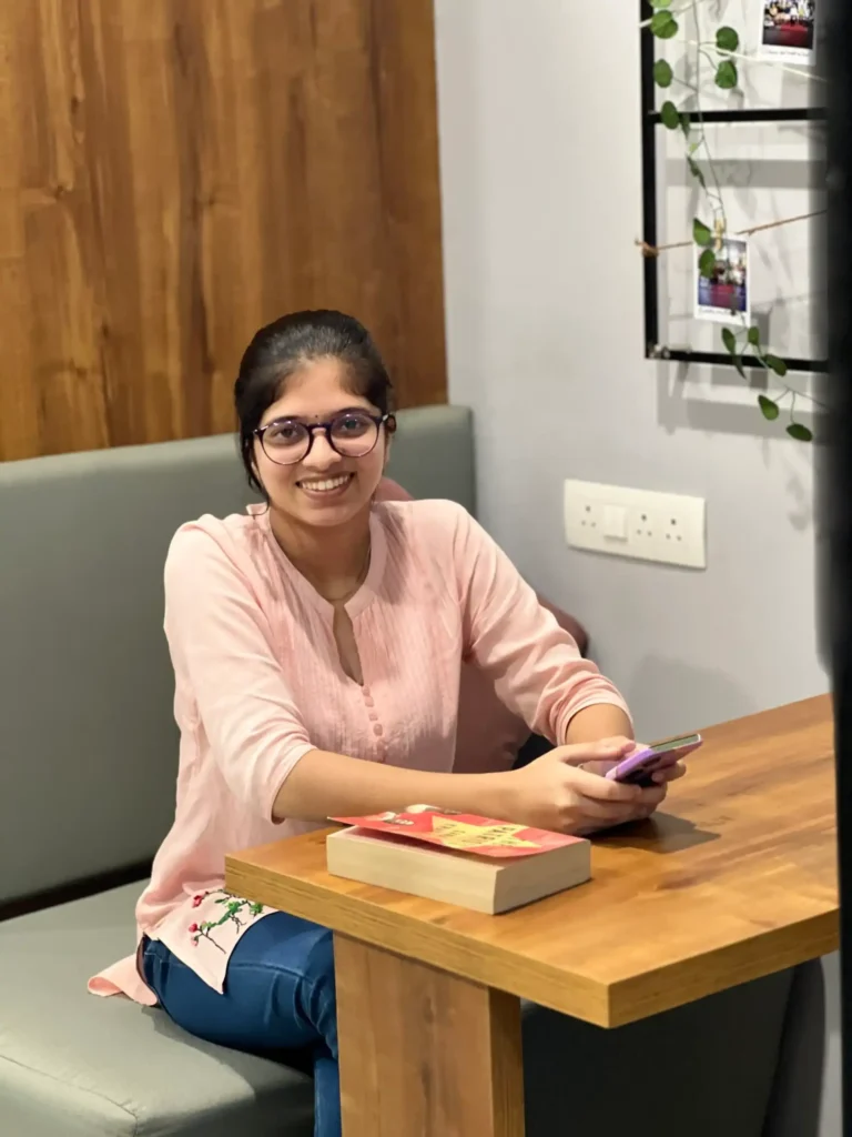 Smiling woman in a light pink kurta and blue pants sits at a cozy café-style table, holding a smartphone with confidence. A book lies open beside her, hinting at creativity and strategy. Framed photos and green vines decorate the wall behind her, adding charm to the relaxed setting. Capturing the essence of the best digital marketing freelancer in Idukki—poised, approachable, and ready to craft campaigns that connect.